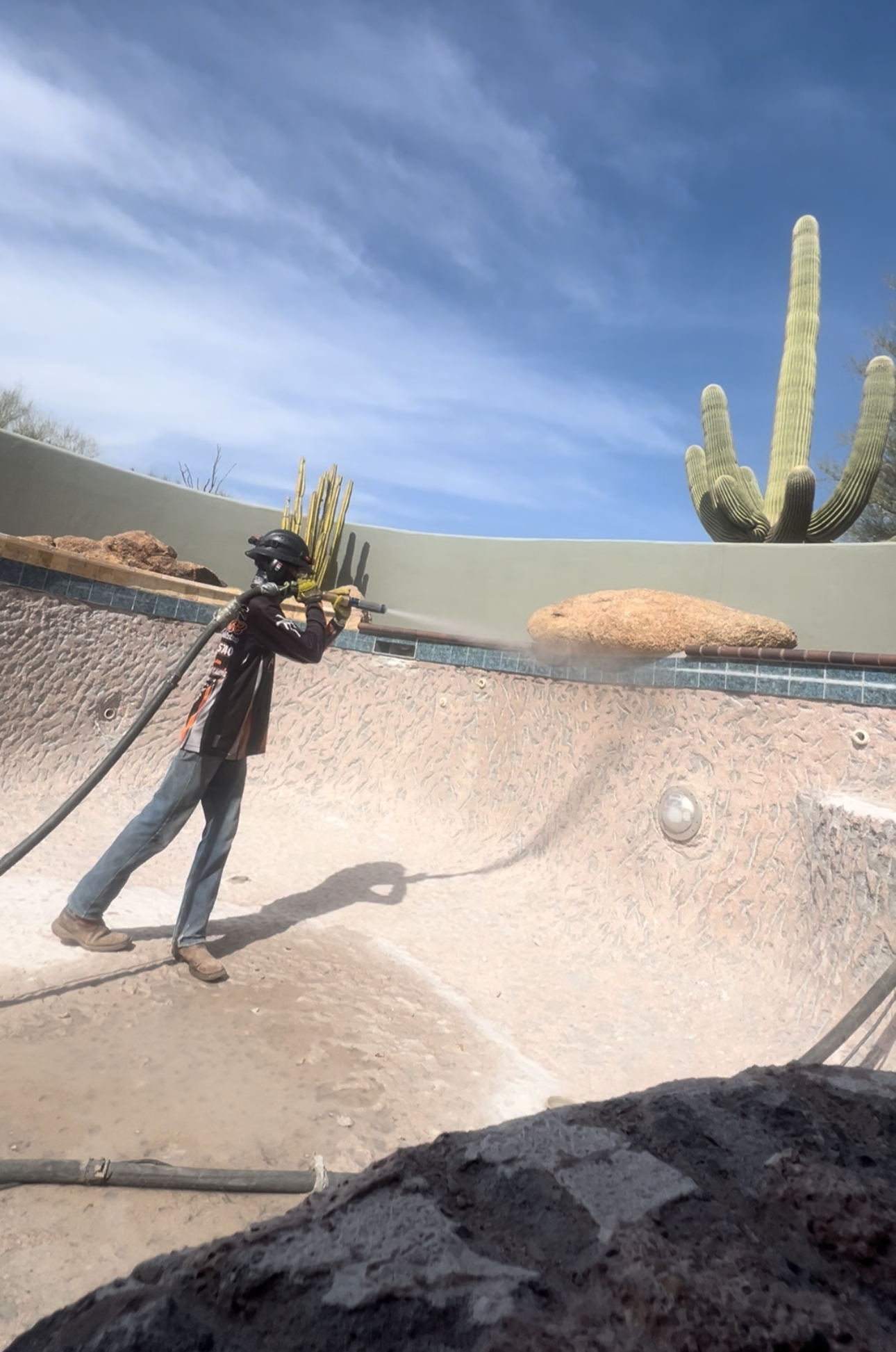 Man sandblasting an empty swimming pool under a blue sky, saguaro cactus in the background.