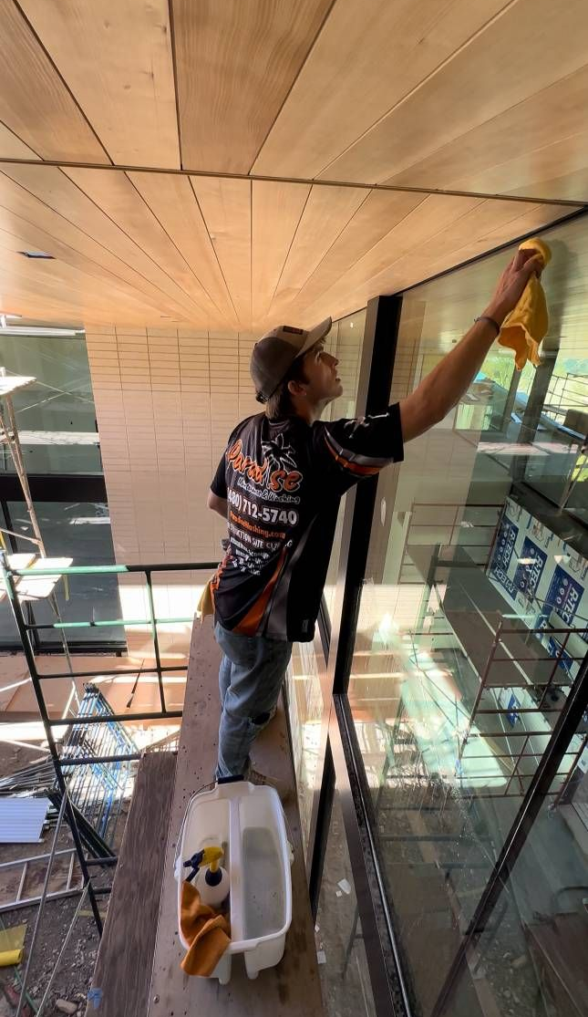 Person on a platform cleaning windows.  They wear a cap and patterned shirt. A bucket sits at their feet.  Exterior shot.