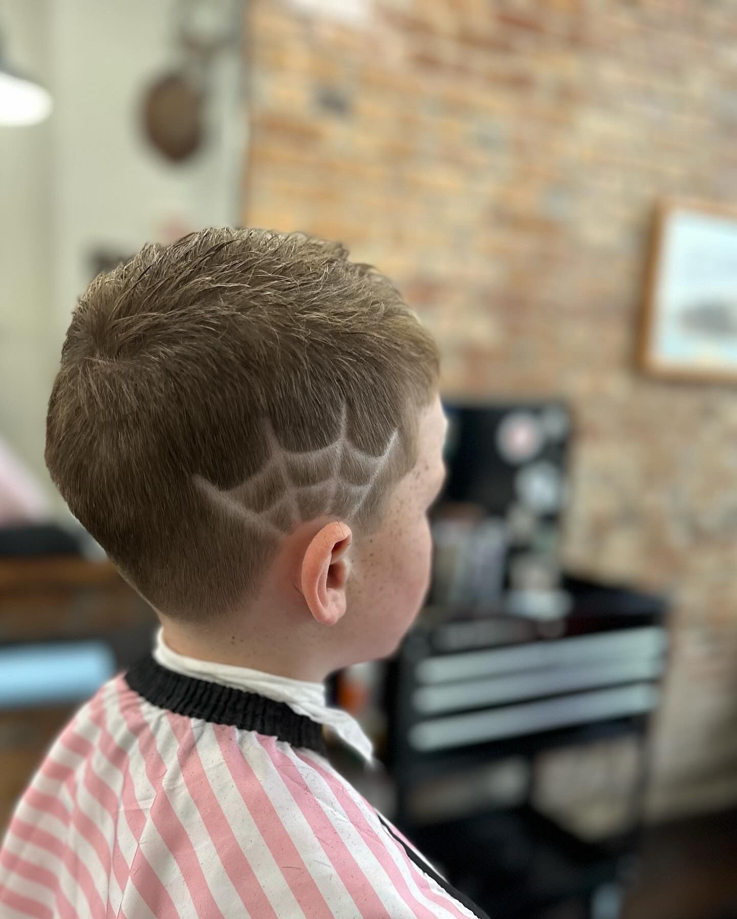 Child with a haircut featuring a spiderweb design on the side of the head, inside a barber shop.