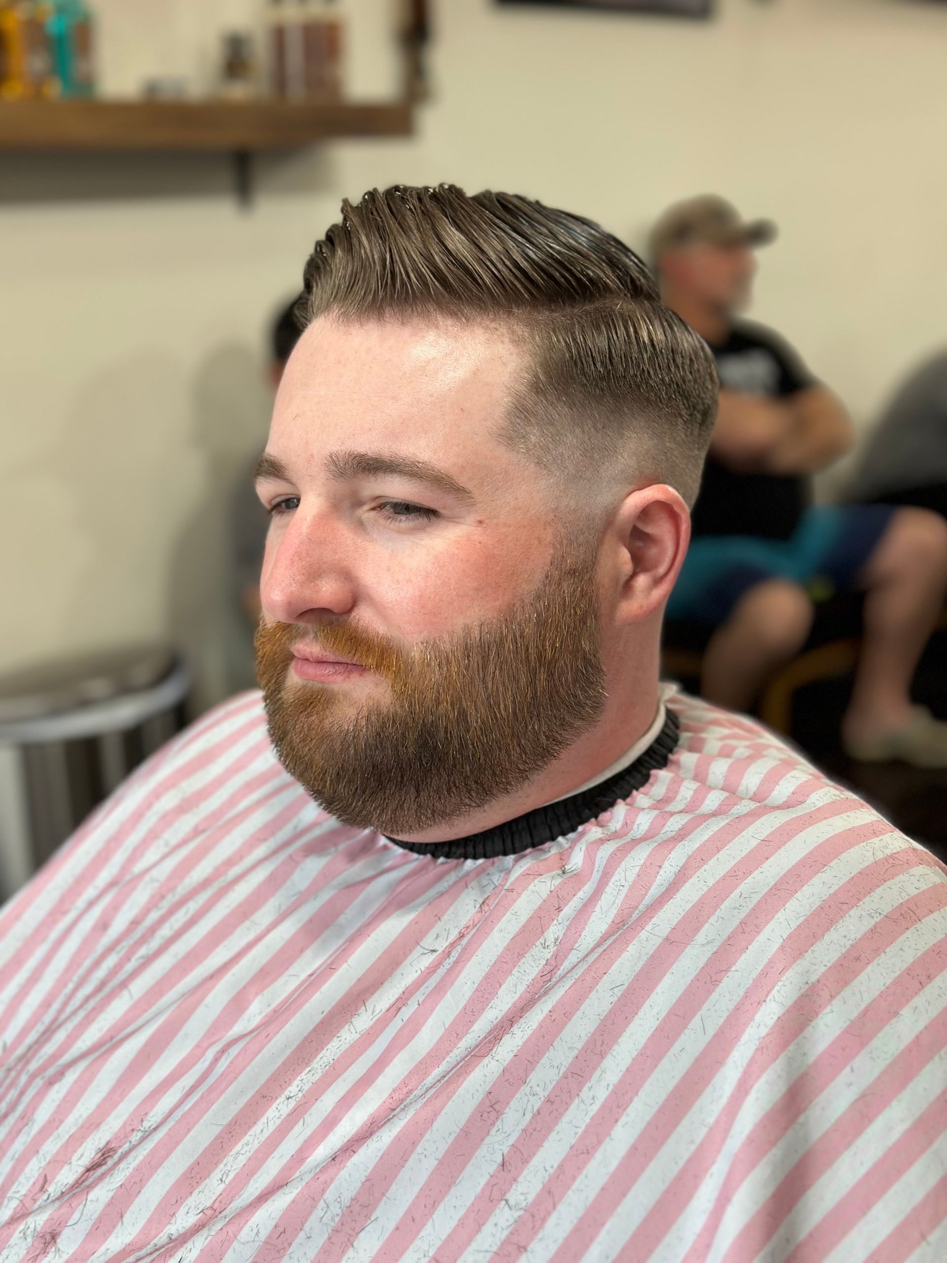 Man with a beard and a faded haircut at a barber shop, wearing a striped barber cape.