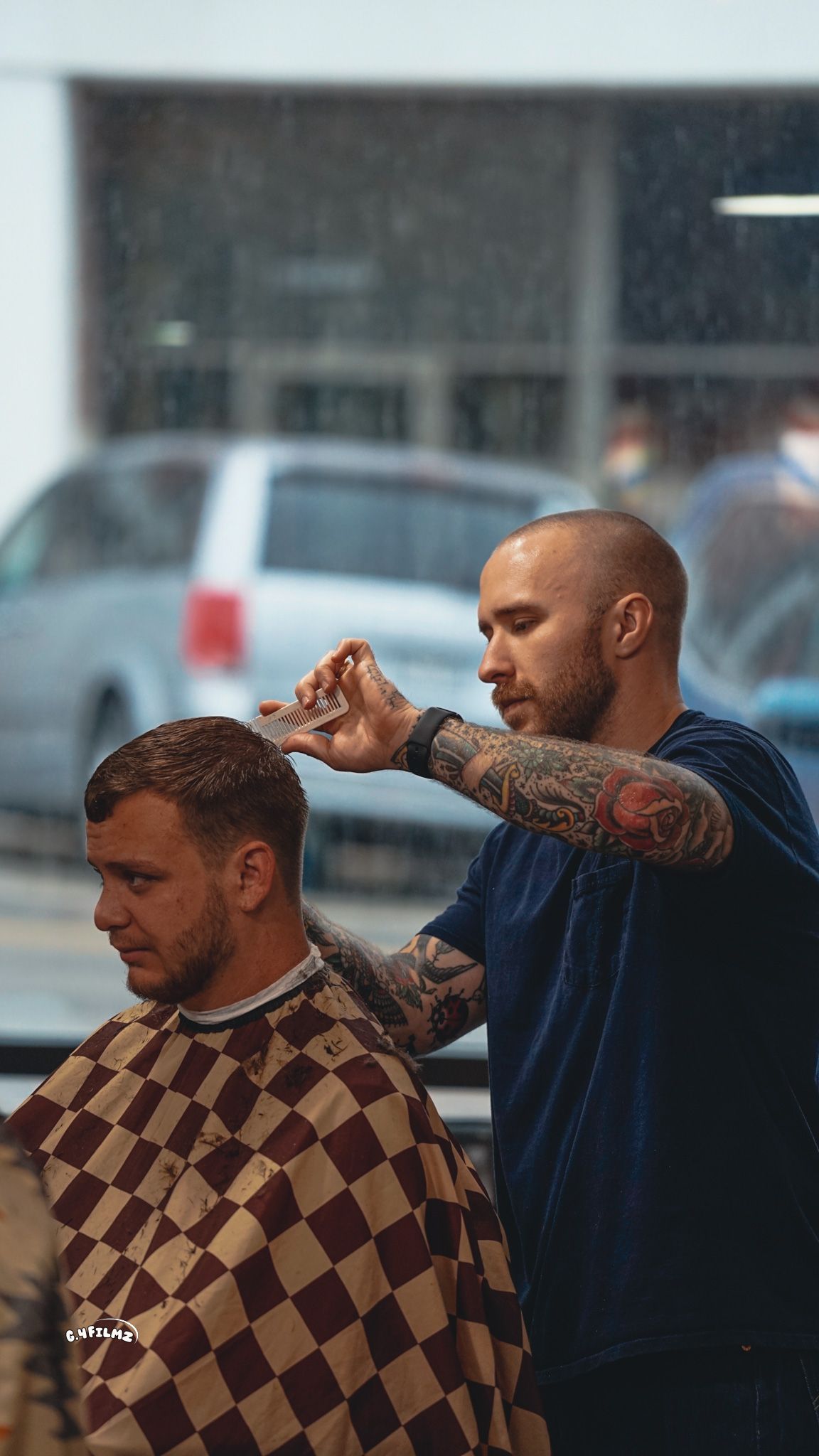 Barber cutting client's hair in shop. Barber has arm tattoos and client wears a checkered cape. Window in background.