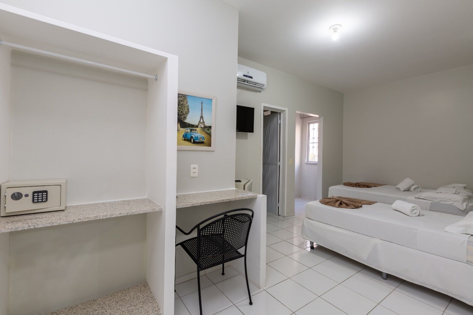 Um quarto de hotel luminoso com duas camas de solteiro, uma escrivaninha com uma cadeira preta, um armário aberto com cofre e piso de azulejos brancos.
