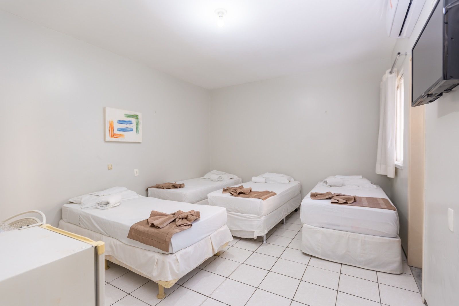 Um quarto de hotel com três camas de solteiro com lençóis brancos e peseiras bege, sobre um piso de azulejos brancos com uma TV instalada na parede.