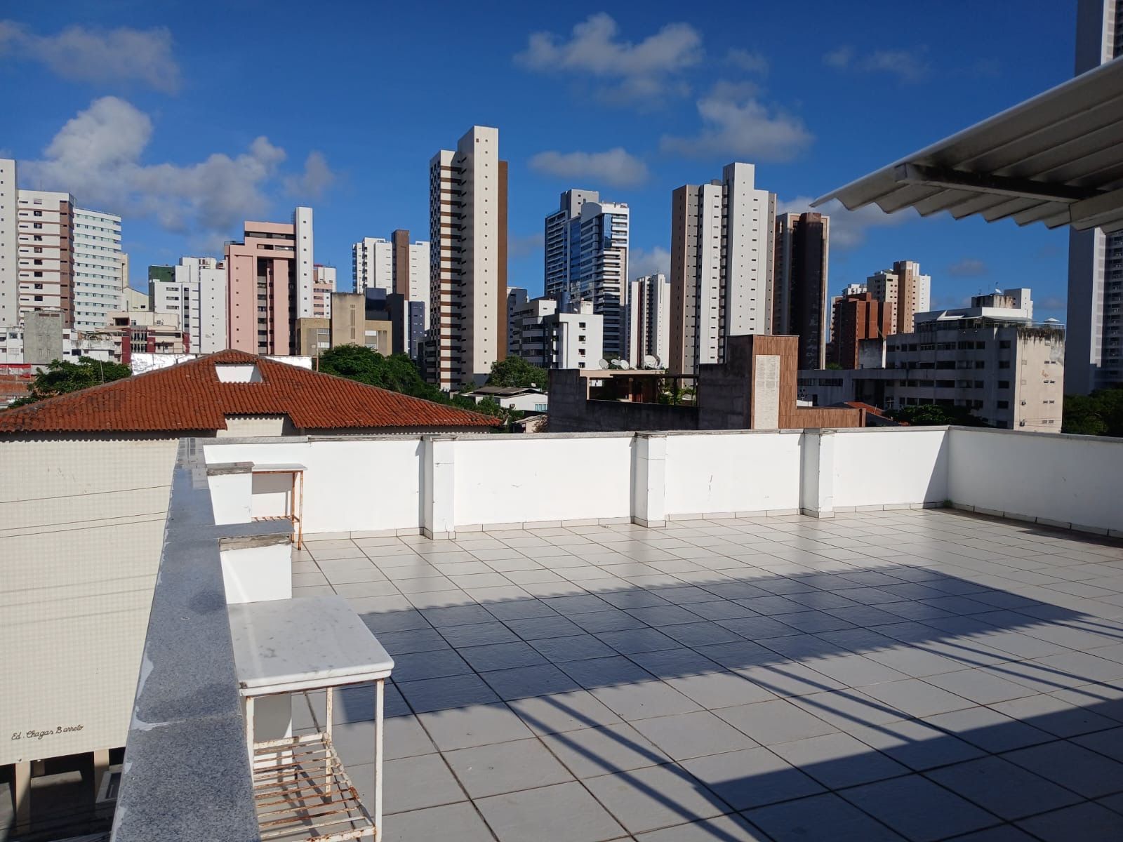 Vista de um terraço na cobertura com azulejos, com vista para o horizonte da cidade, com prédios altos sob um céu azul com nuvens brancas.