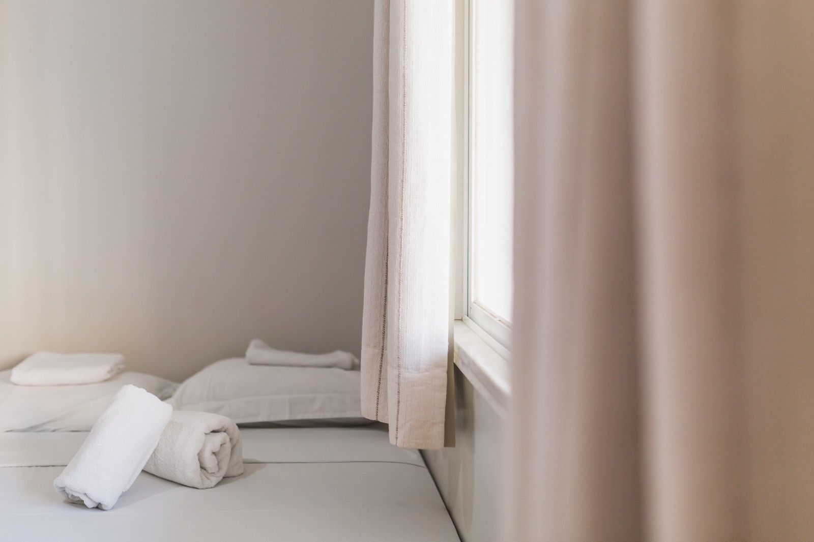 Duas toalhas brancas enroladas estão sobre uma cama ao lado de travesseiros, perto de uma janela com cortinas claras em um quarto de tons neutros.