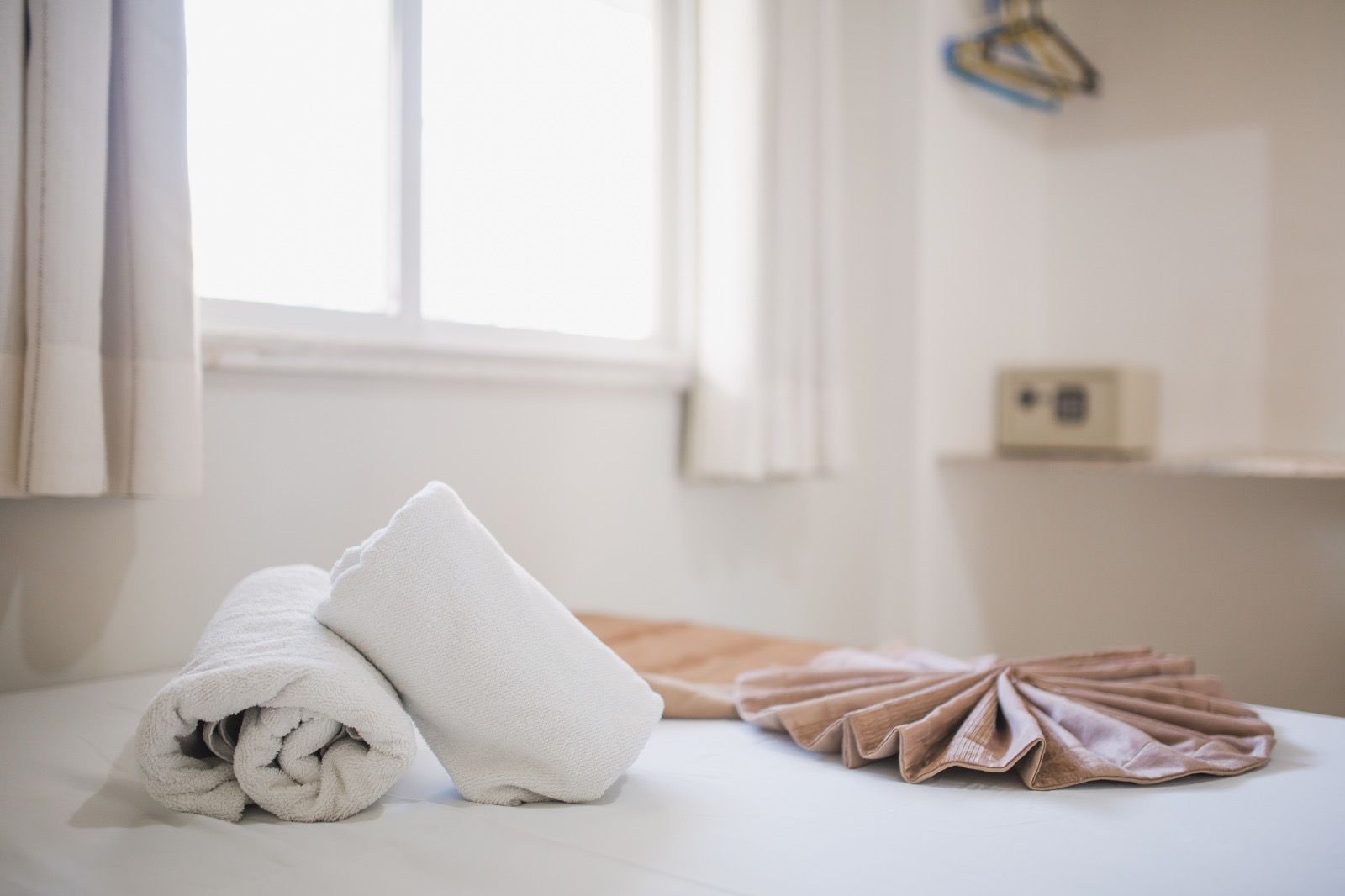 Toalhas brancas enroladas e uma toalha bege dobrada dispostas sobre uma cama em um quarto de hotel, com um cofre visível ao fundo.