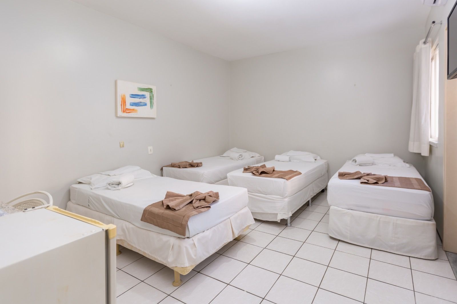 Um quarto de hotel com piso de azulejos brancos, paredes bege e quatro camas de solteiro arrumadas com lençóis brancos e mantas marrons.