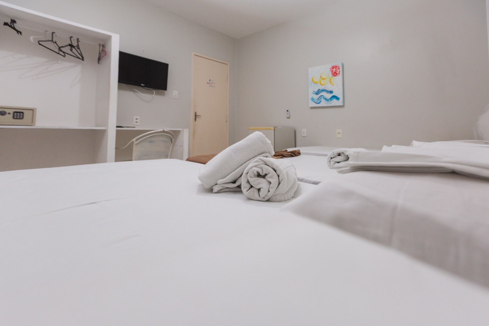 Um quarto de hotel luminoso com uma cama com toalhas brancas dobradas, um armário com cofre, uma escrivaninha e obras de arte em uma parede clara.