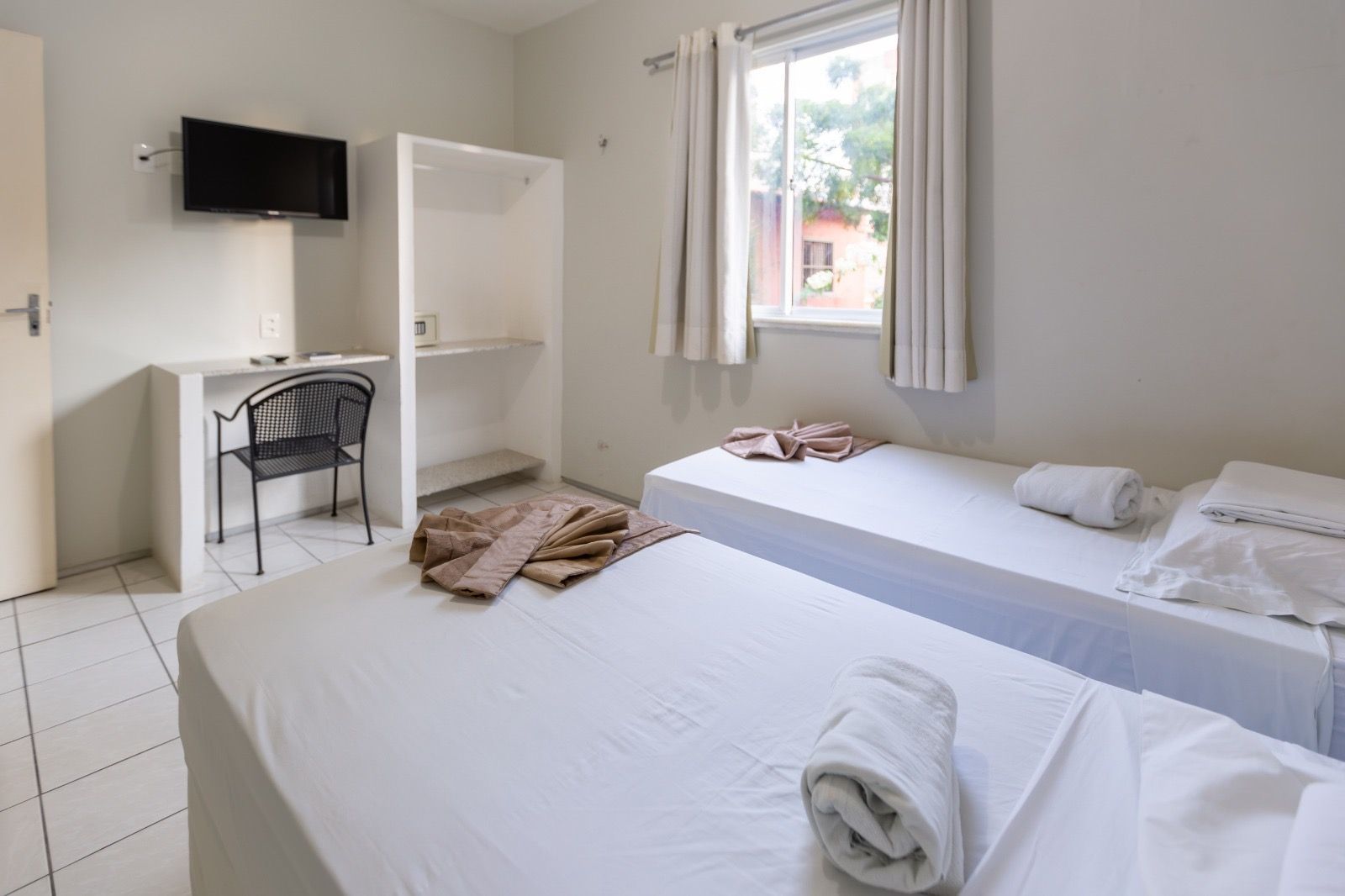 Um quarto de hotel com duas camas de solteiro com lençóis brancos, uma pequena escrivaninha com uma cadeira preta e um guarda-roupa perto da janela.