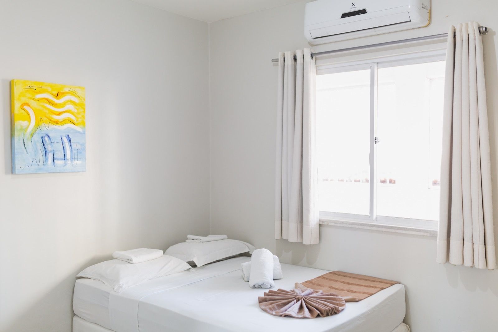 Um quarto de hotel minimalista com uma cama branca impecavelmente arrumada, uma vibrante pintura abstrata em amarelo e azul e uma janela.