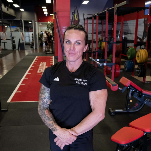 Woman standing in a gym, wearing a black shirt with tattooed arms and red workout equipment behind her