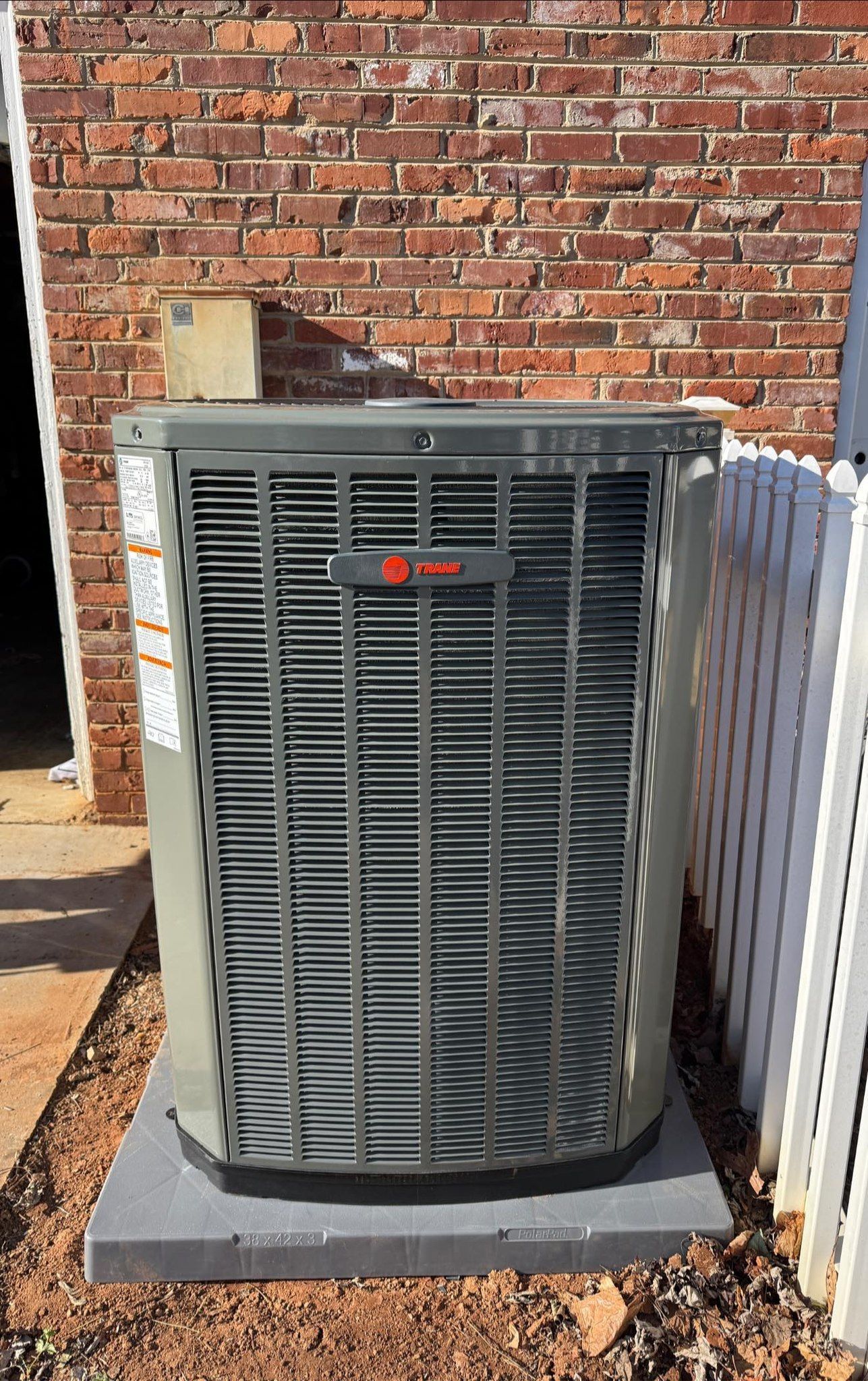 Outdoor air conditioner unit beside a brick wall and white fence on a concrete pad