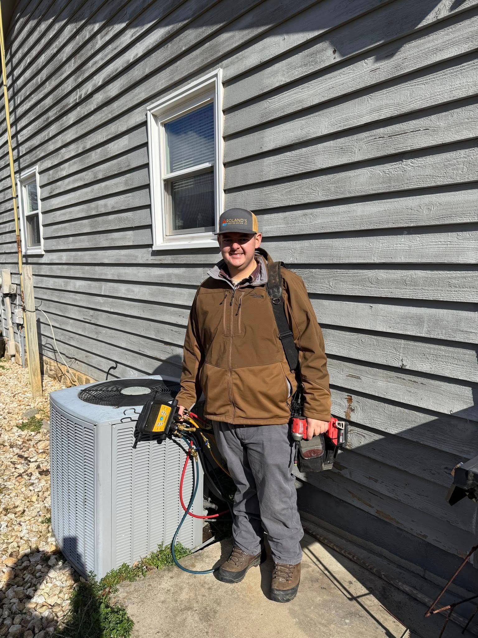 Person in work clothes standing beside a house wall near an outdoor HVAC unit, holding tools.