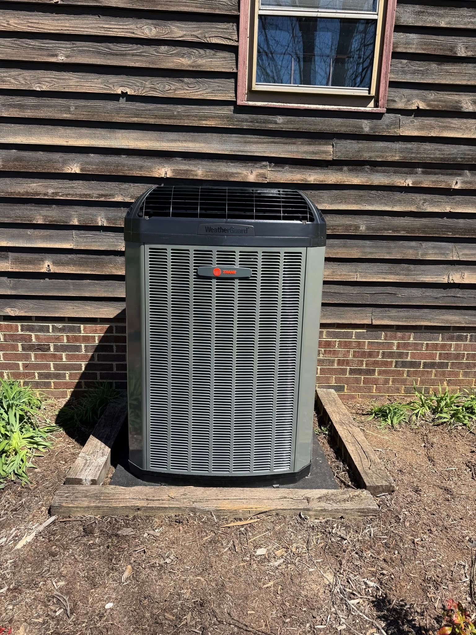 Outdoor central air conditioner unit beside a house with dark wood siding and a small window above it