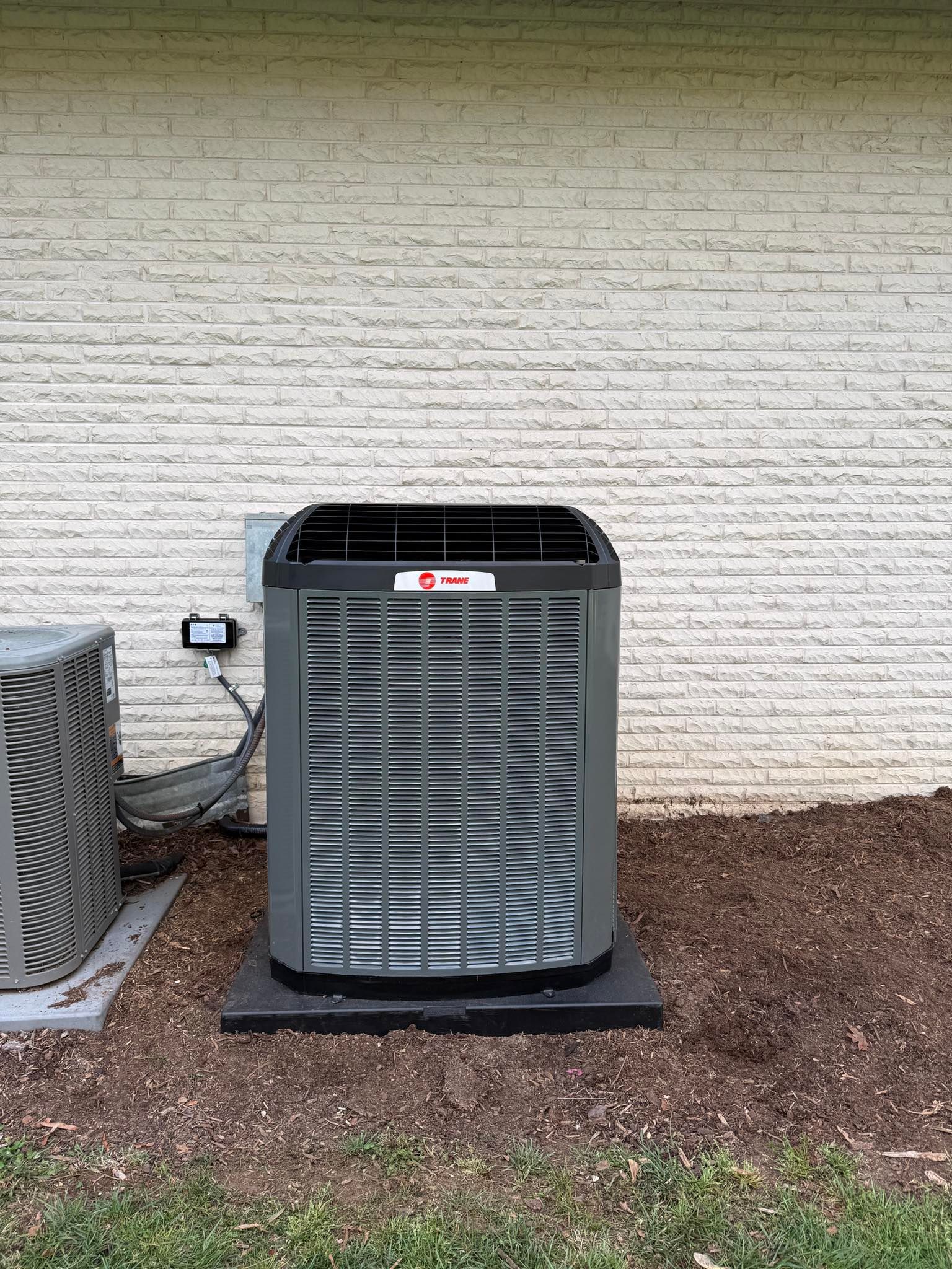 Outdoor air conditioner unit beside a brick wall, with a second unit partially visible on the left.