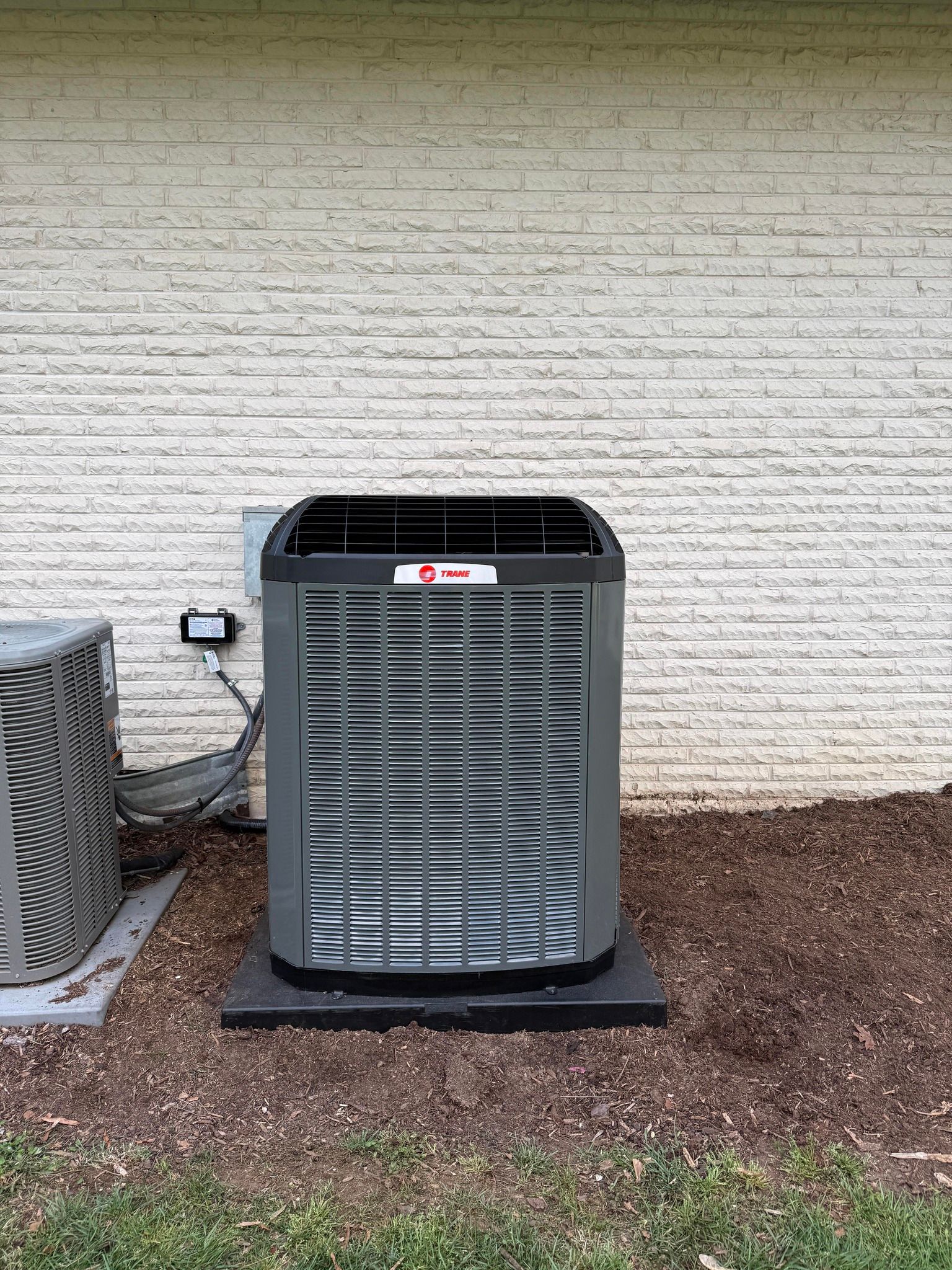 Outdoor HVAC unit beside a brick wall, with a smaller fan unit on the left and mulch around it.