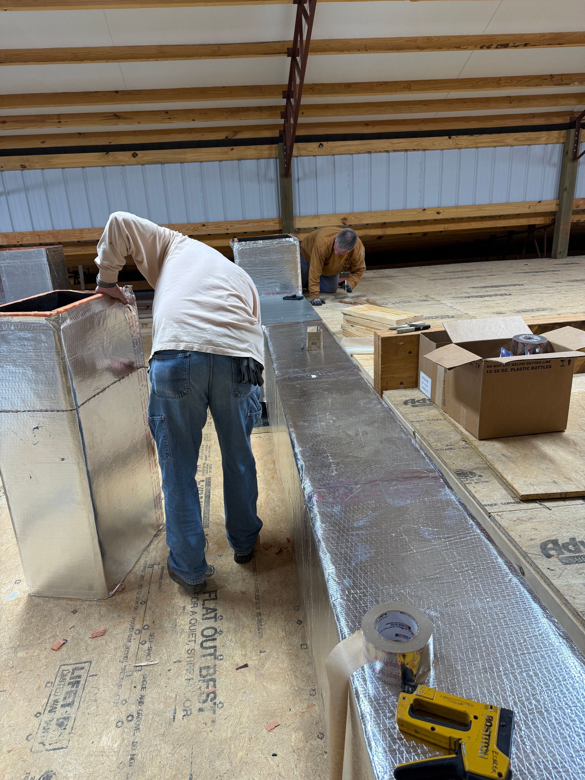 Worker in a warehouse bending over a long metal workbench with tools and boxes nearby.