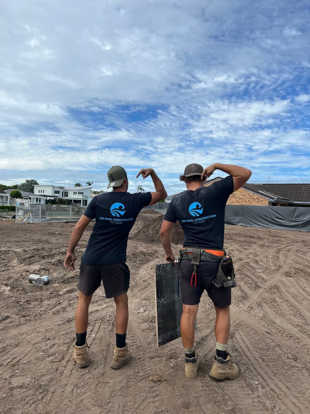 Two workers in black shirts stand on a muddy construction site under a cloudy sky, surveying the area.