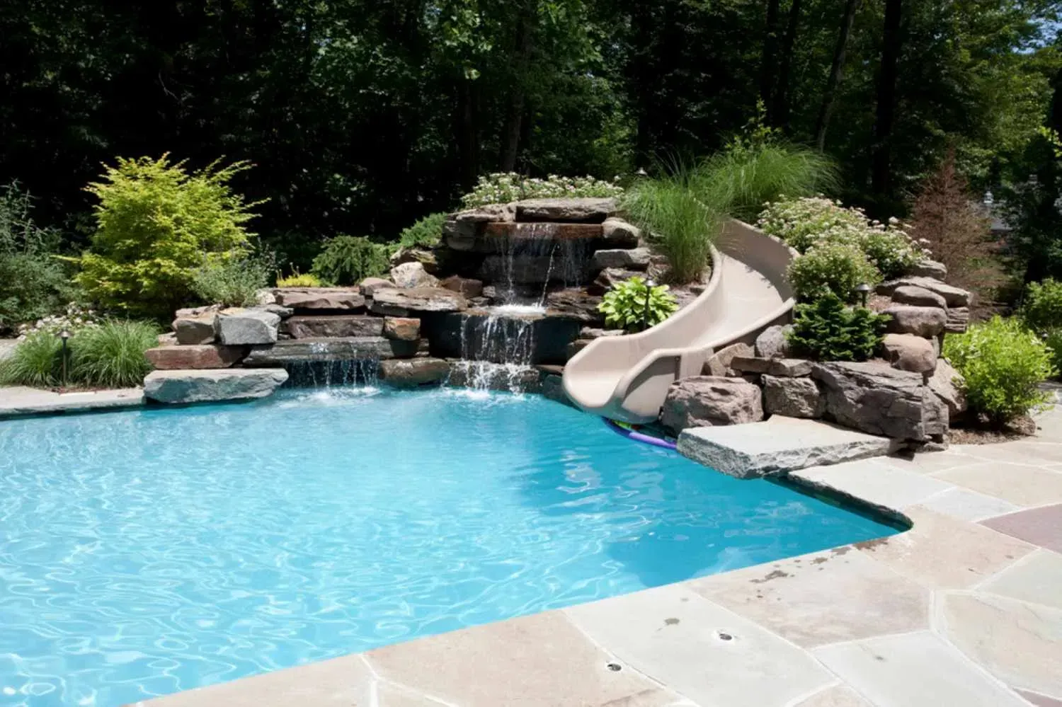 Backyard pool with rock waterfall, slide, and turquoise water surrounded by greenery