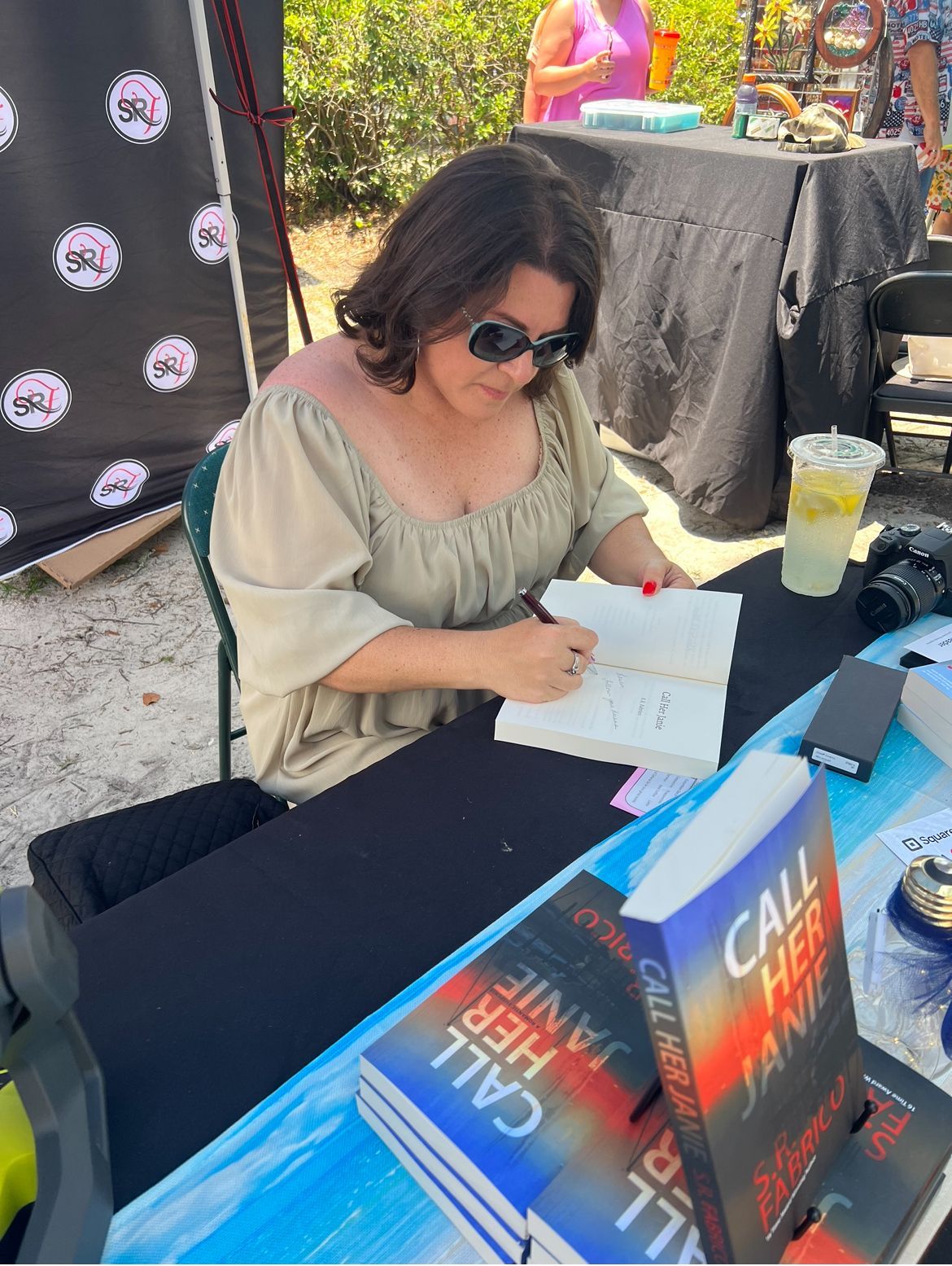 A woman is sitting at a table signing a book called call her jane