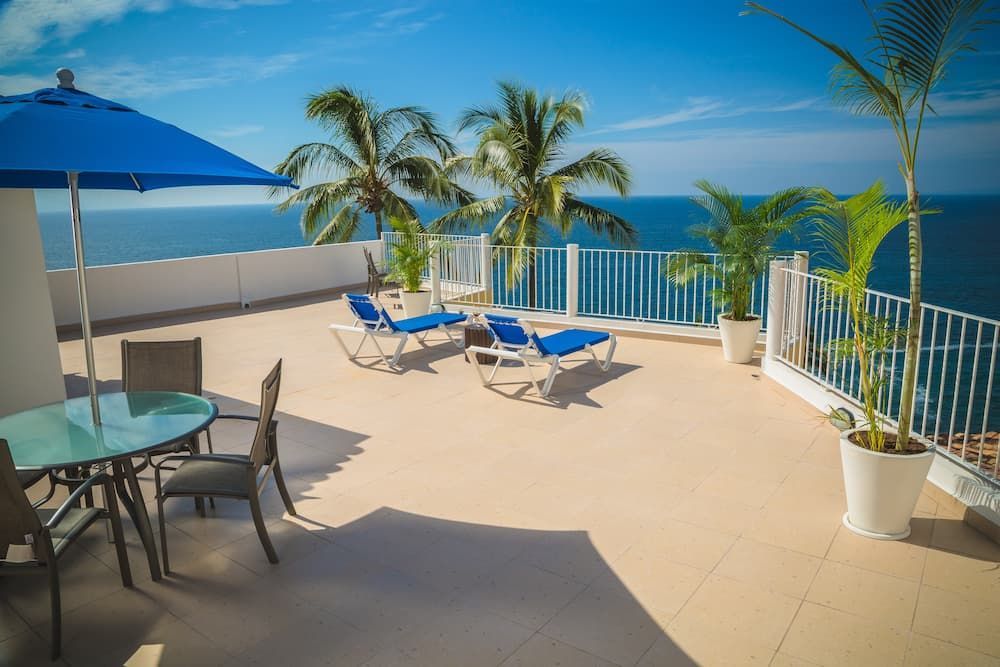 Rooftop patio with ocean view; blue umbrella, lounge chairs, table, palm trees, white railing, and blue sky. — Unique Waterproofing Solutions in North Rocks, NSW