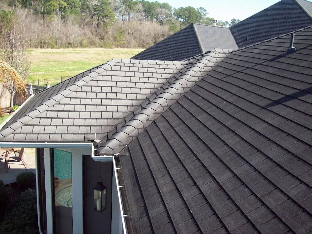 Dark gray shingle roof of a house with a gutter and a backdrop of trees and a field.