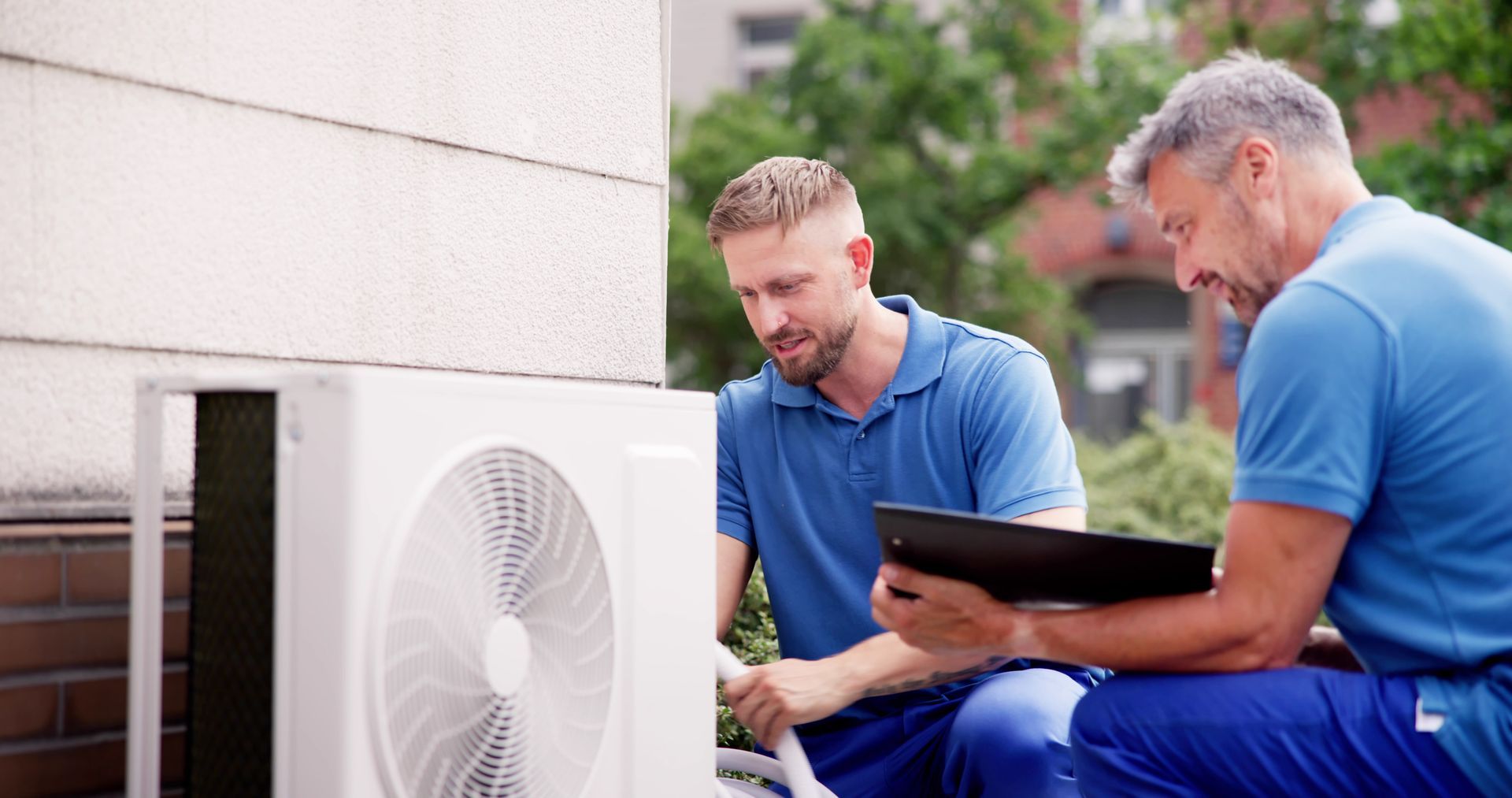 Two men are working on an air conditioner outside of a building.