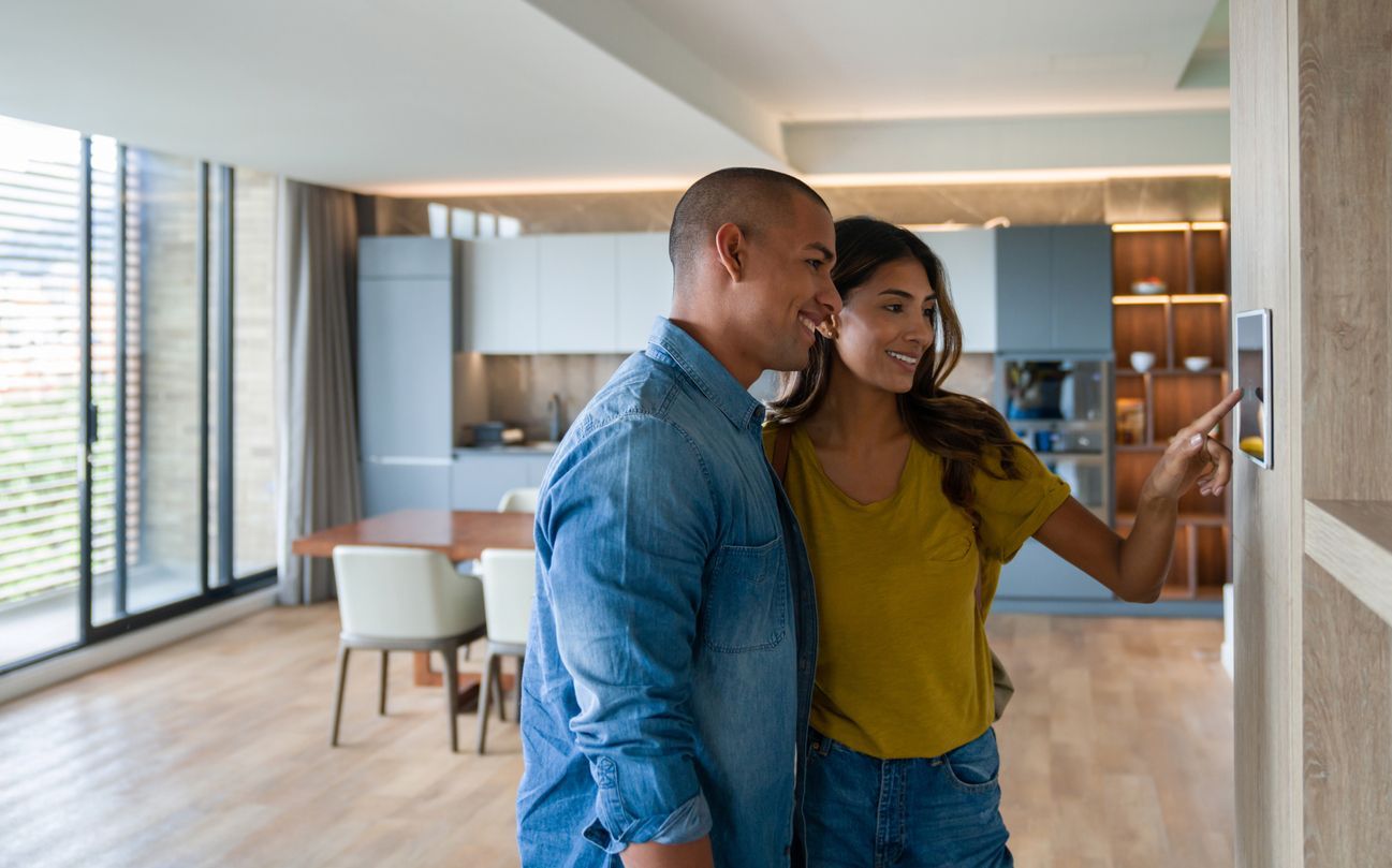 A man and a woman are standing in a living room looking at a thermostat.