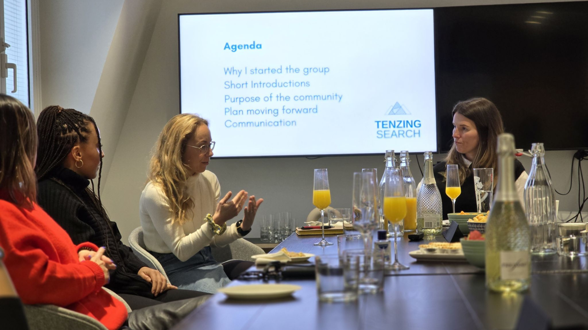 Four people sit around a table at a meeting, facing a screen showing an agenda with the Heading "TEAZING SEARCH".