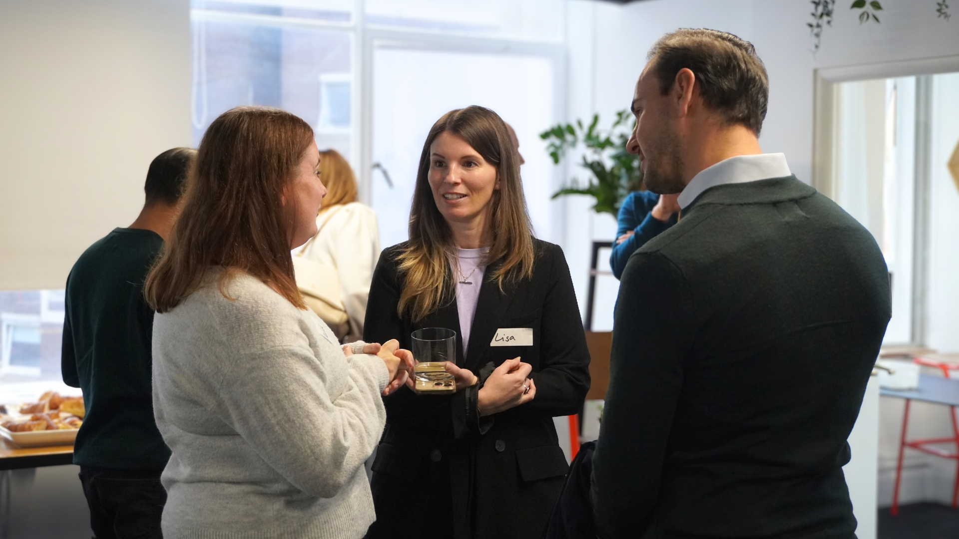 Three people standing in a bright office, talking and holding drinks during a social gathering.