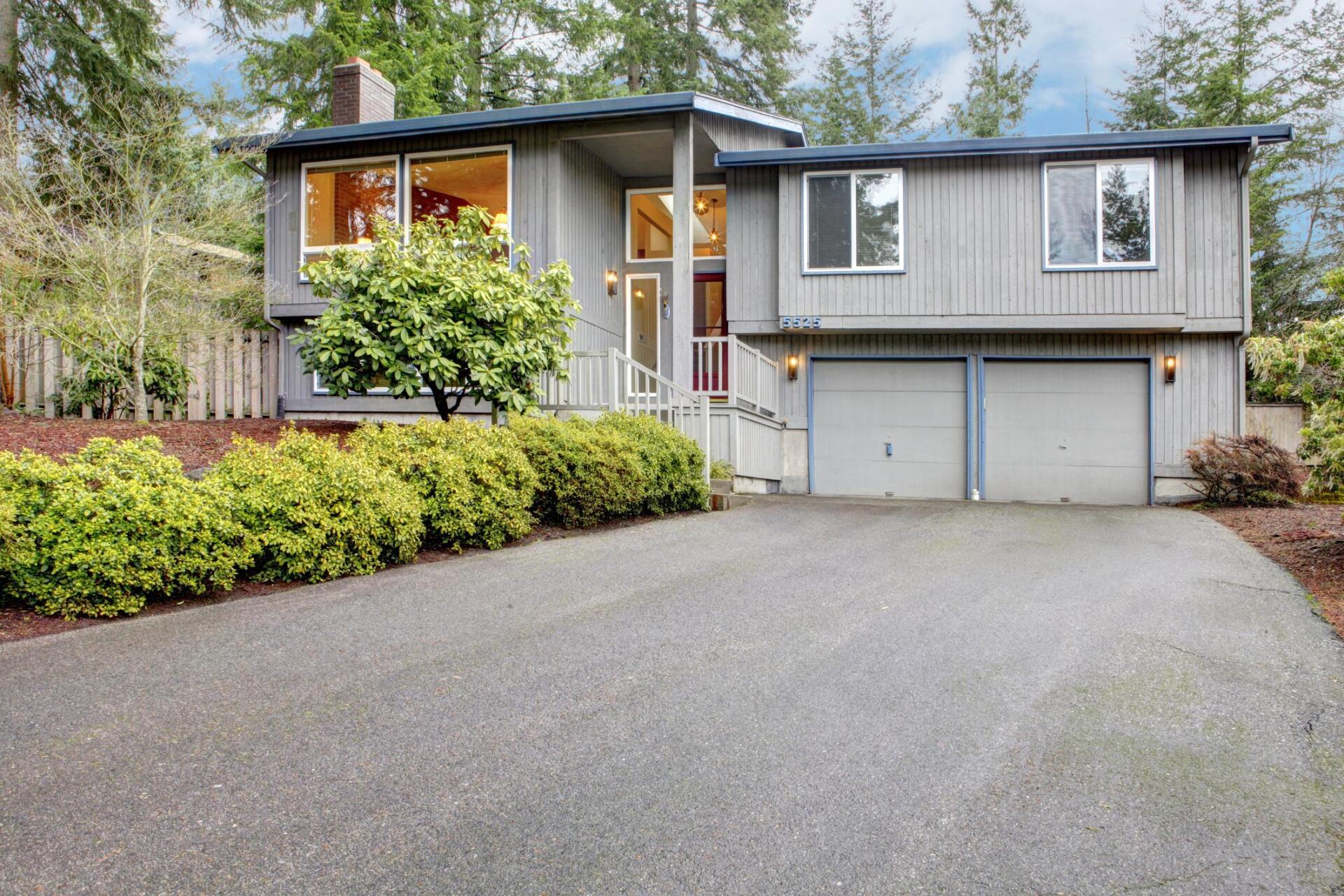 A split-level grey house with a two-car garage, large windows, and a manicured hedge along a paved driveway.