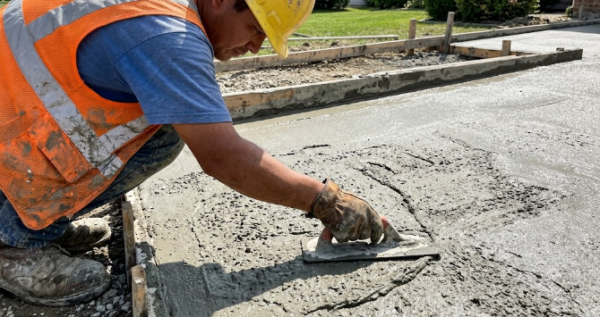 Close up of a professional construction worker wearing safety gear, using a metal trowel to expertly smooth out fresh wet concrete on a new residential driveway project.
