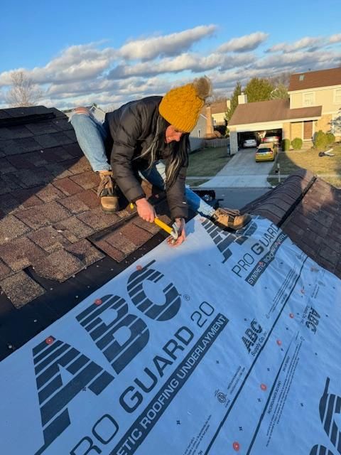 Worker in orange beanie installing roofing underlayment on a shingled roof at sunset