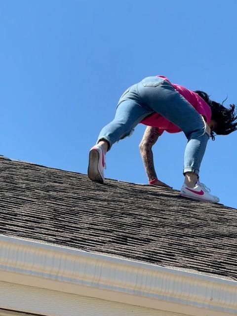 Person kneeling on a shingled roof against a blue sky, inspecting or working near the roof edge.