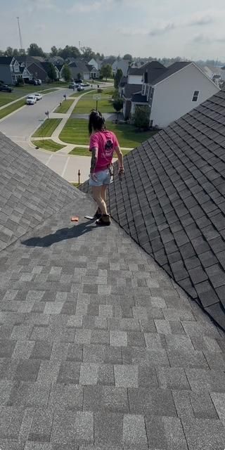 Person walking down a shingled rooftop in a suburban neighborhood, wearing a pink shirt and shorts