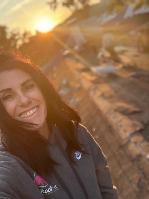 Smiling woman in a black jacket standing by a sunlit beach at sunset
