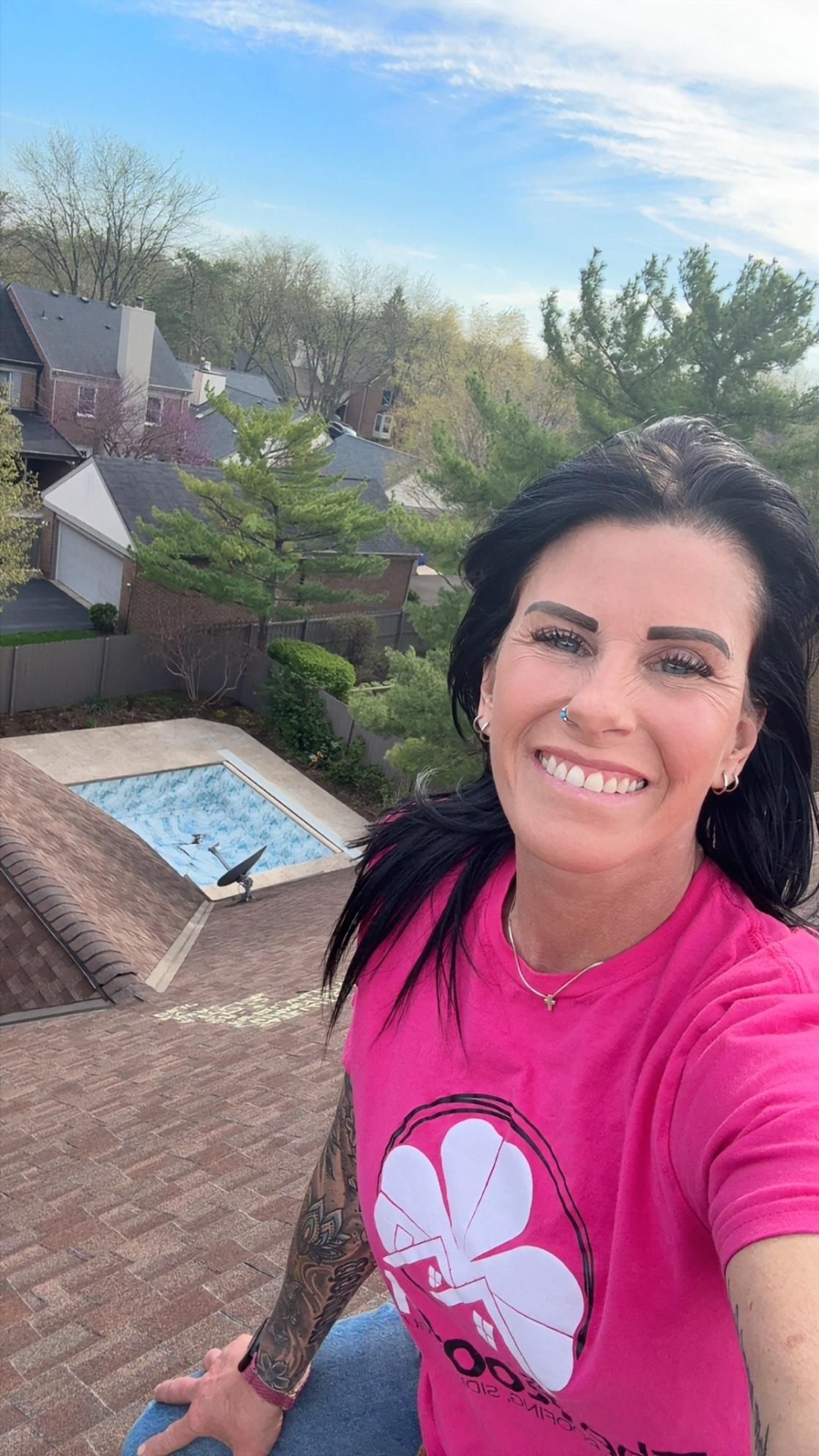 Smiling woman in pink shirt taking a selfie on a rooftop, with a backyard pool and trees below.