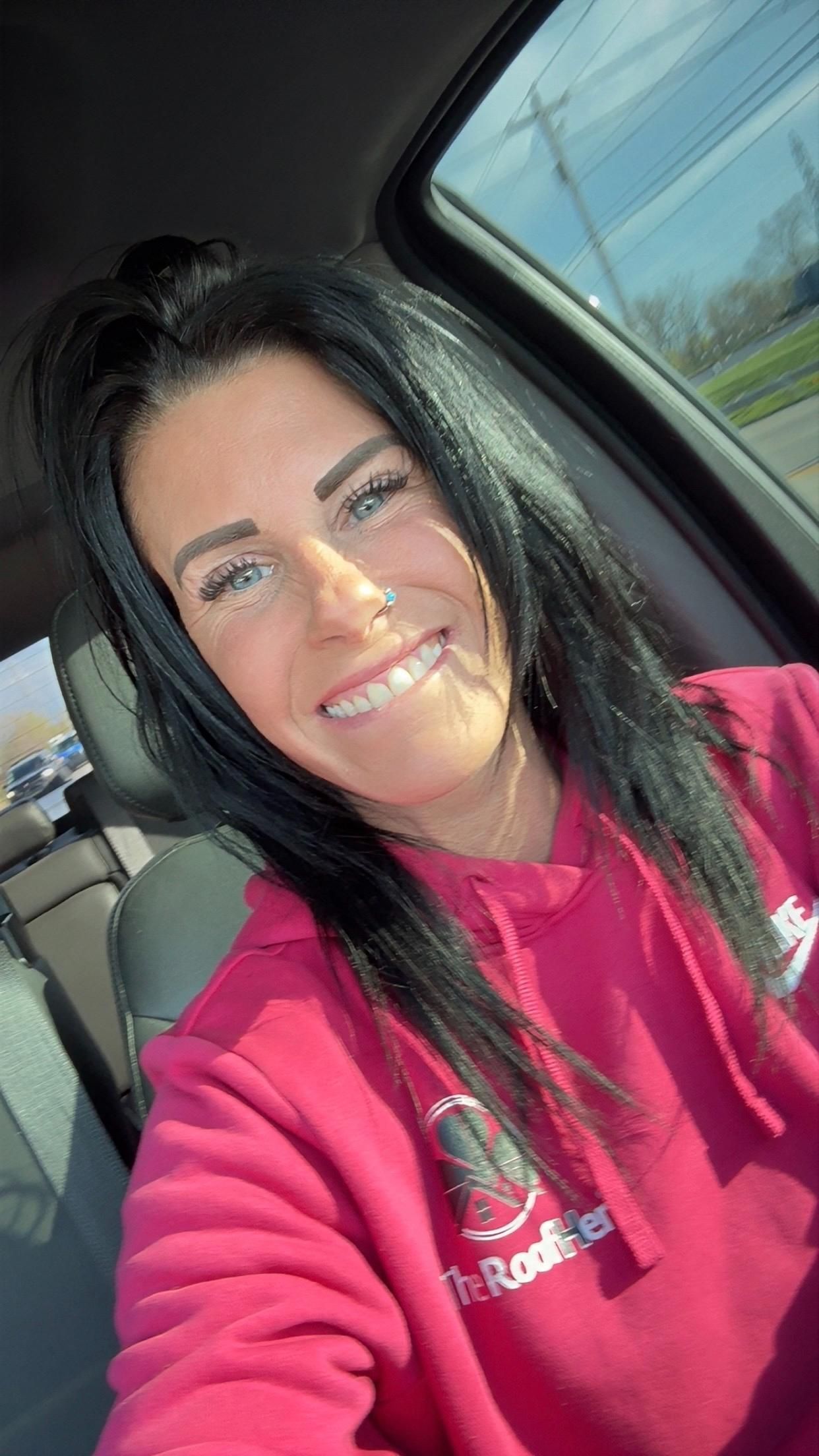 Woman smiling in a car, wearing a bright pink sweatshirt in sunlight.
