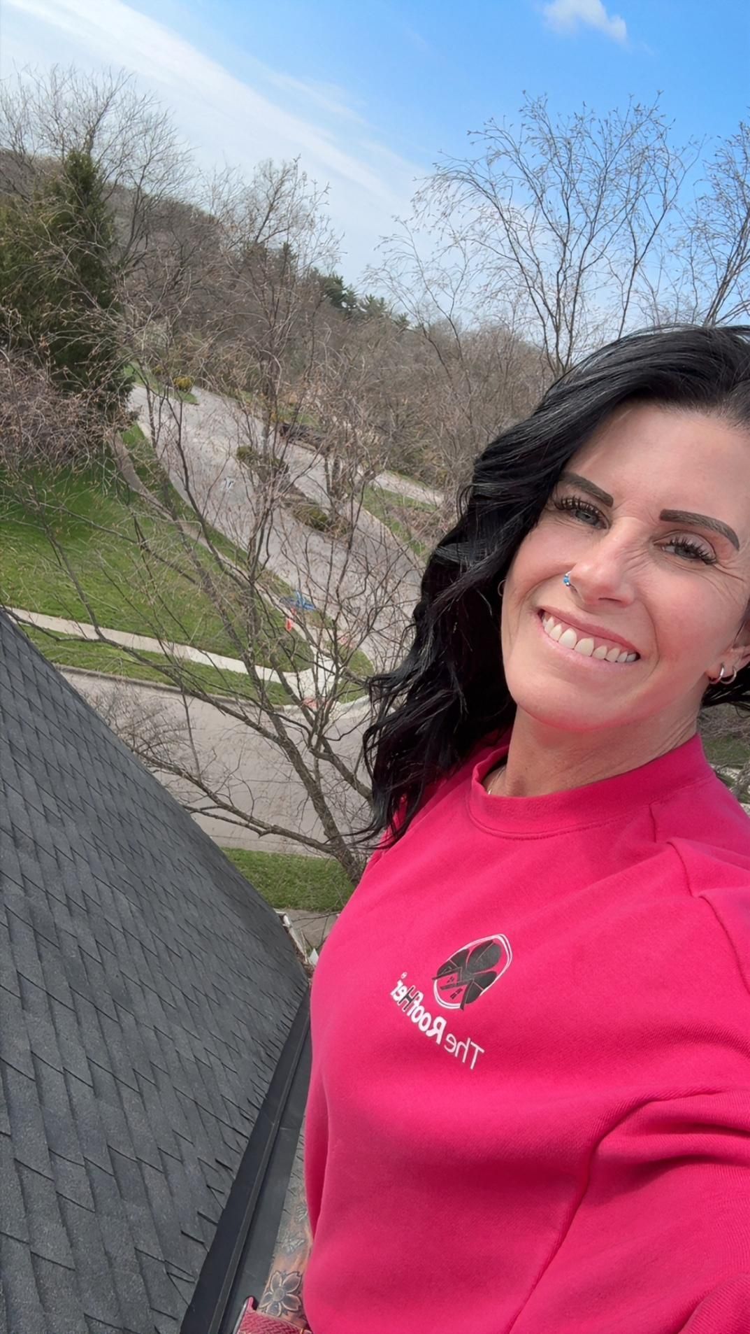 Woman in a bright pink sweatshirt smiling outdoors beside a rooftop with trees and blue sky behind her