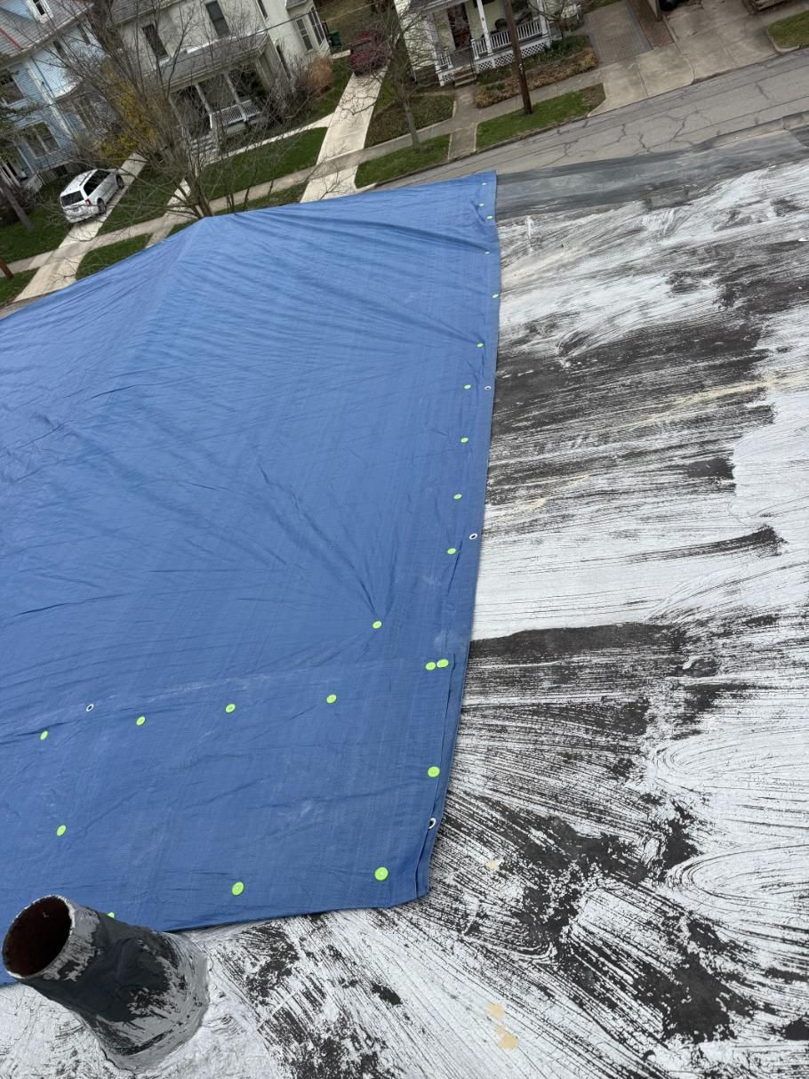 Blue tarp on a weathered flat rooftop with a vent pipe and nearby houses in the background