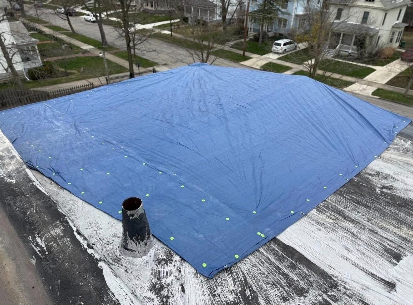 Blue tarp covering a flat rooftop, secured with weights, with a neighborhood street and houses in the background