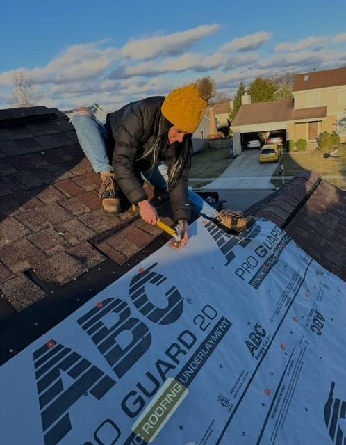 Worker in yellow beanie installing roof underlayment on a shingled rooftop on a sunny day