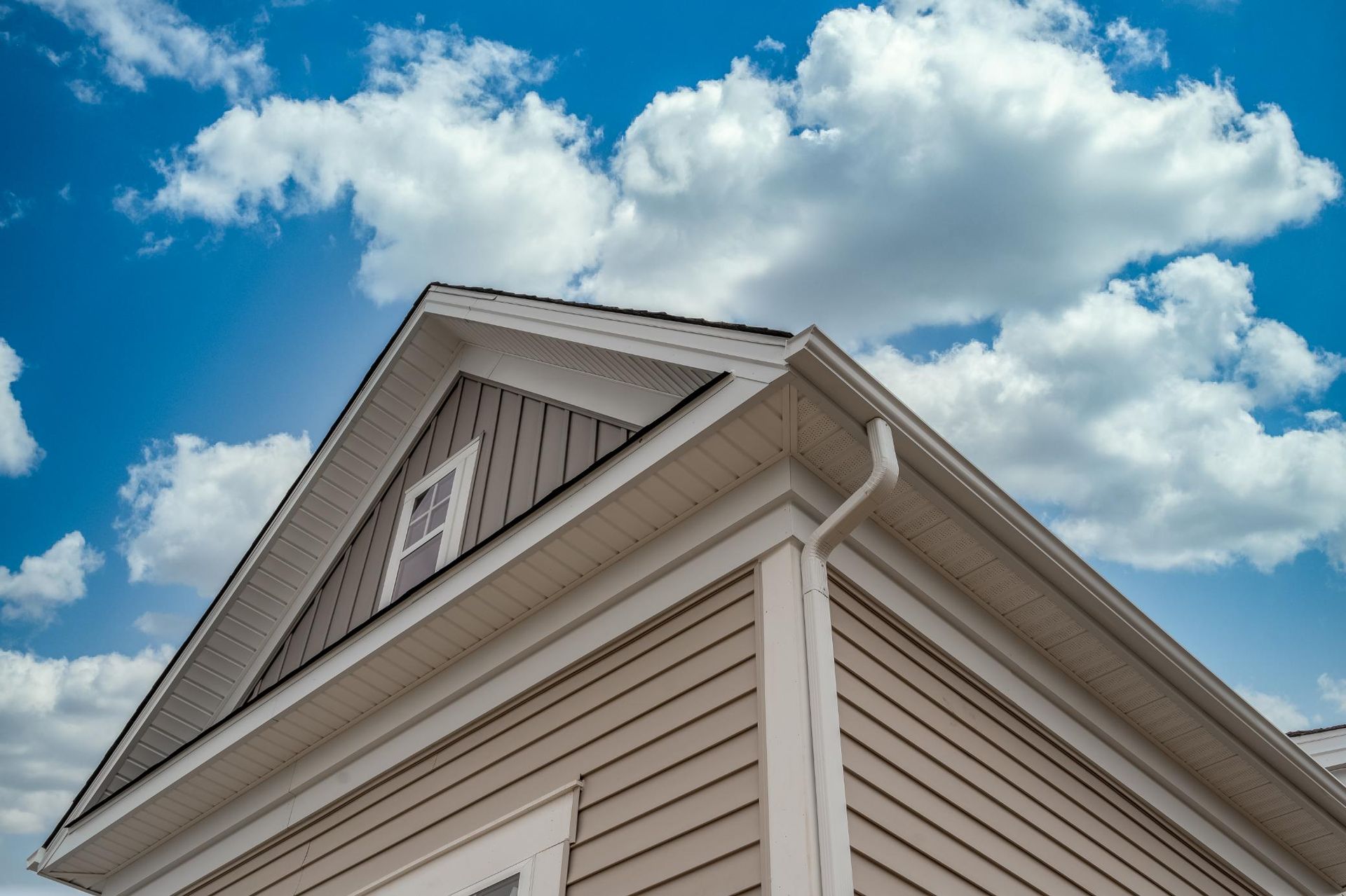 Beige house corner with white trim and gabled roof against a blue sky with clouds
