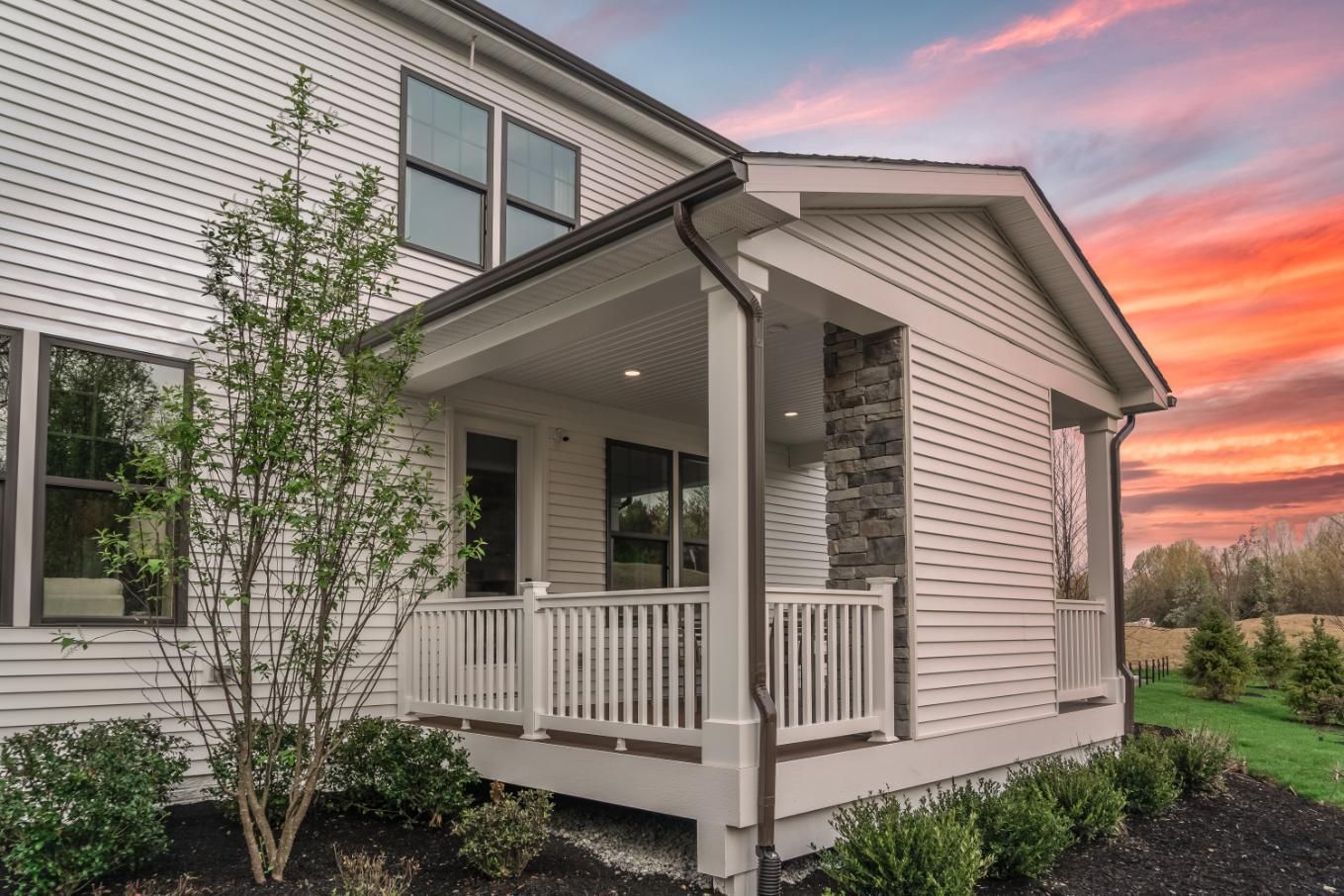 Suburban house porch with white railing at sunset, framed by greenery and a pink-orange sky