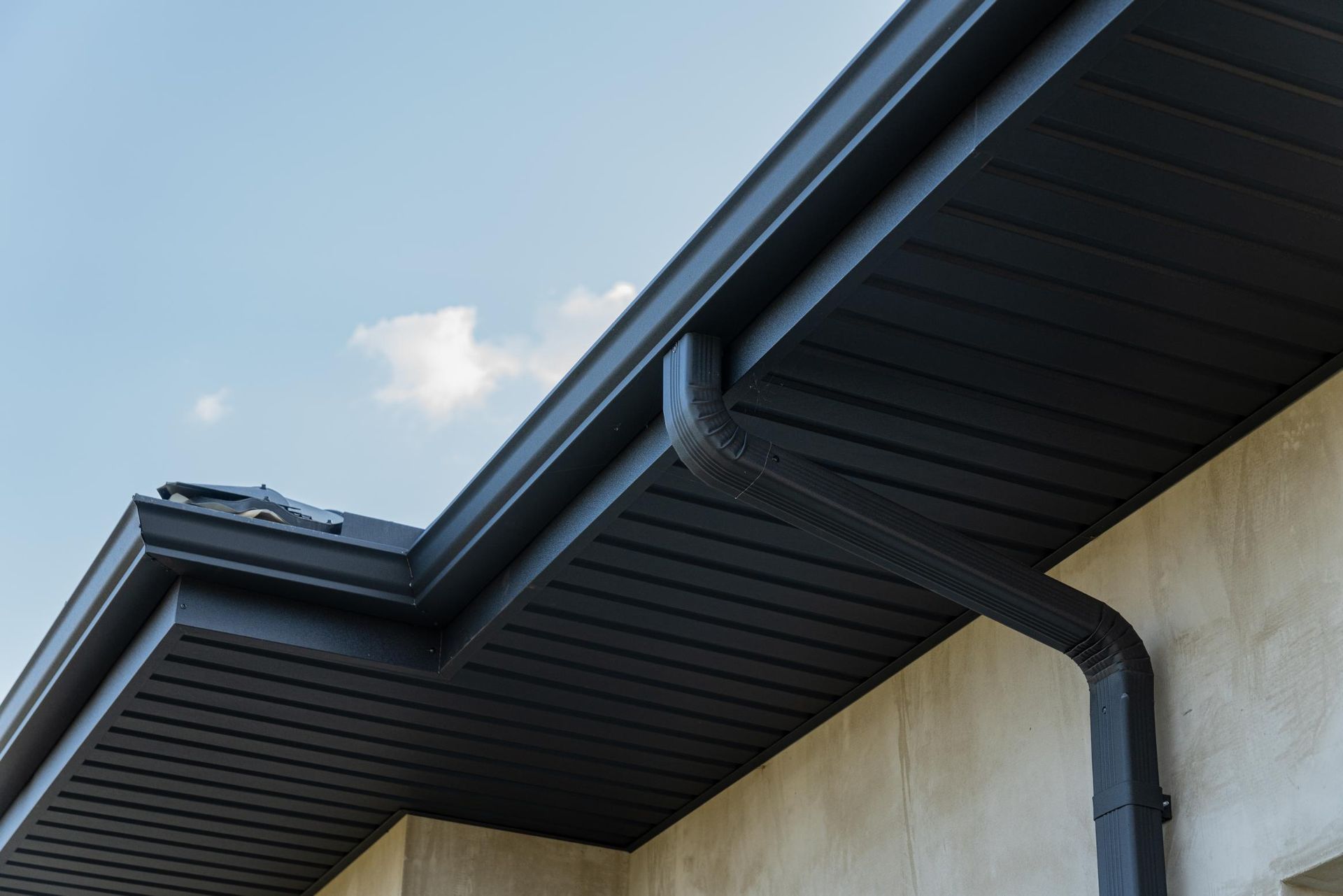 Close-up of a house roof with dark gutters and downspout against a blue sky