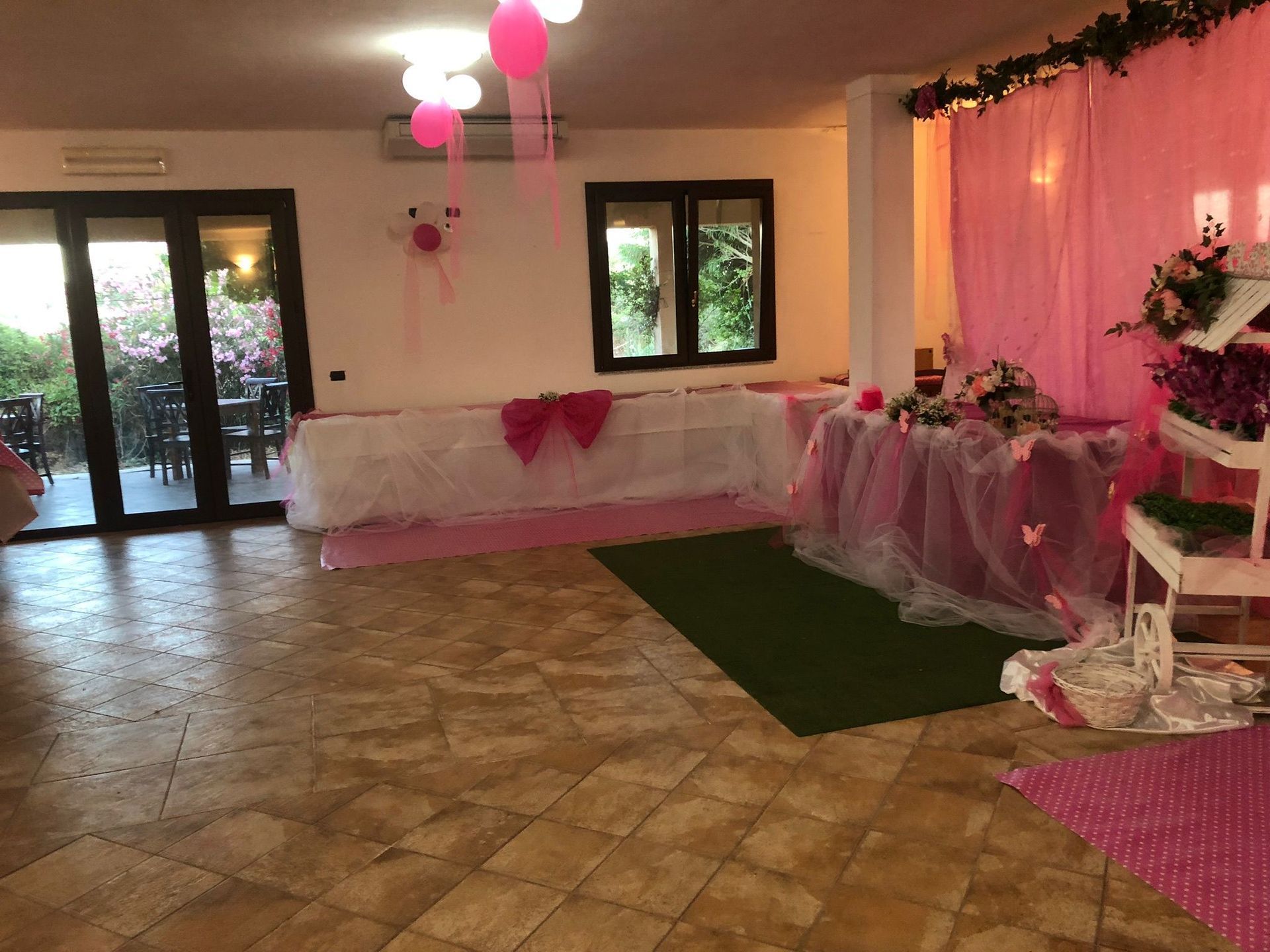 Sala per feste decorata con palloncini rosa e bianchi, decorazioni per la tavola e un tappeto verde.