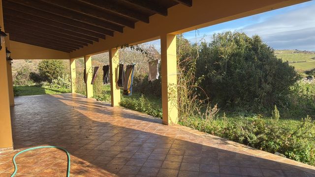Patio coperto con pavimento piastrellato, travi e colonne in legno, affacciato su un paesaggio verde e soleggiato.