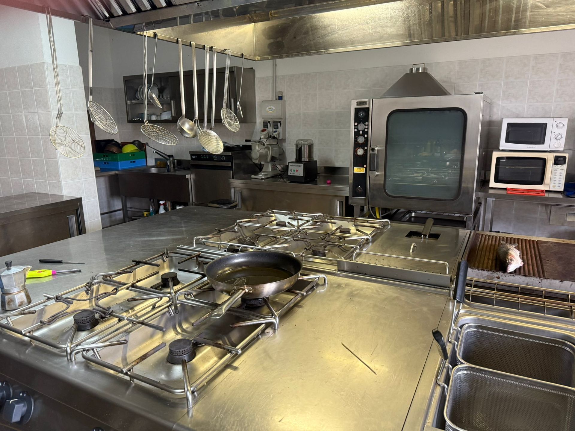 Cucina professionale con piano cottura in acciaio inox, utensili appesi e un televisore retrò sullo sfondo.