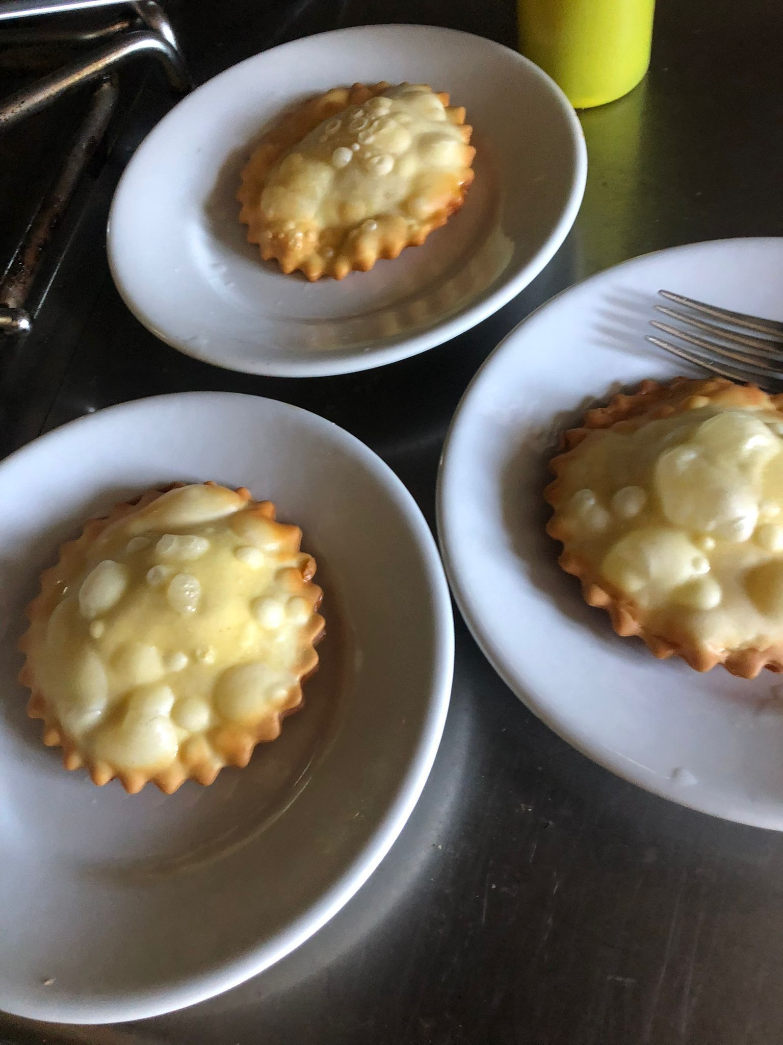 Tre piccole crostatine di frutta ricoperte di glassa bianca, disposte su piatti bianchi e appoggiate su un tavolo di metallo.