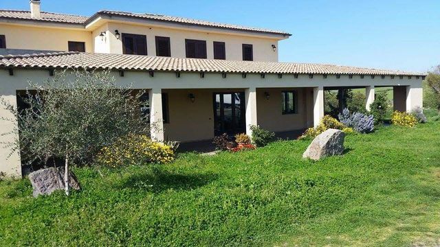 Casa di campagna con veranda coperta, pareti color beige e prato verde con rocce e arbusti.